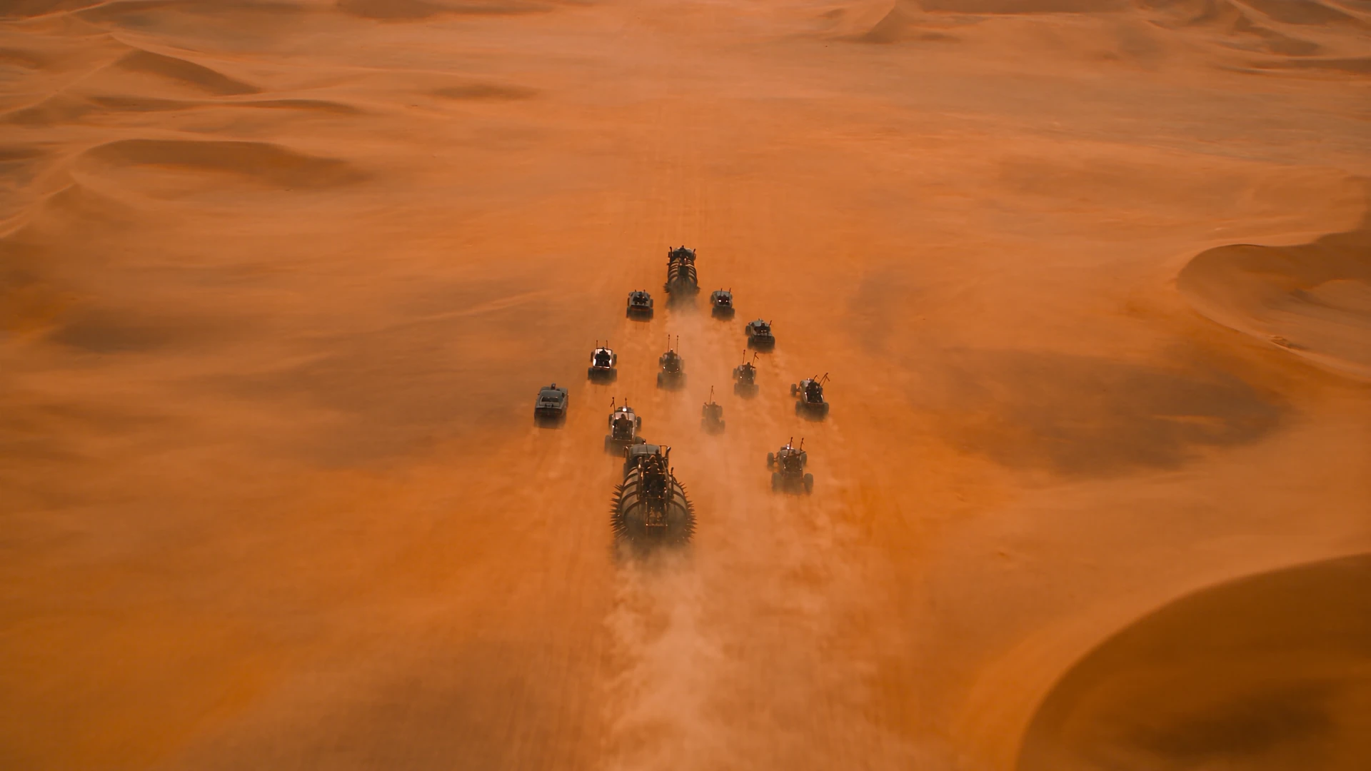 Aerial view of a convoy of post-apocalyptic war vehicles driving in formation across vast orange sand dunes, kicking up dust trails