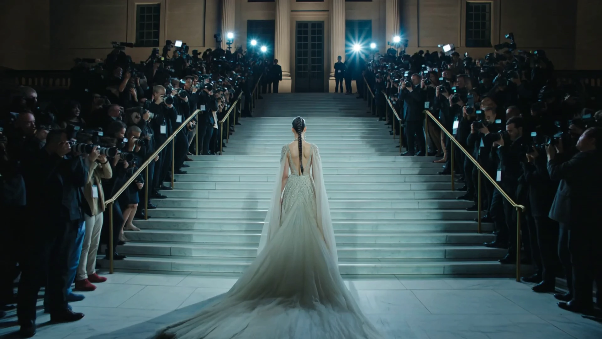 Woman in an elaborate white gown with a long train climbing grand stone steps at night, surrounded by rows of photographers on both sides