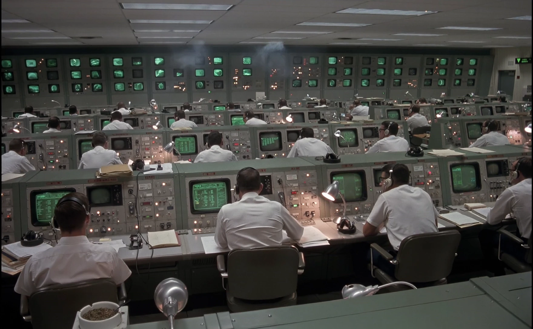 Operators working at consoles in a retro mission control room filled with green monitors.